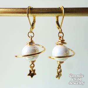 May include: A pair of gold-tone earrings. Each earring has a white, iridescent sphere encircled by a gold wire, with a gold star charm. The earrings hang from gold lever-back clasps. The text "Cosmic Child Jewelry" is visible.
