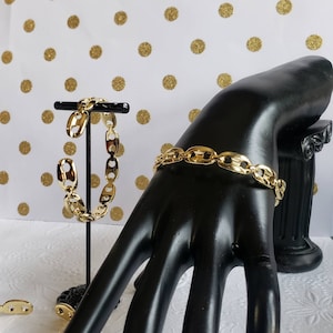May include: Two gold chain bracelets with a mariner link design. One bracelet is on a display stand and the other is on a black mannequin hand.