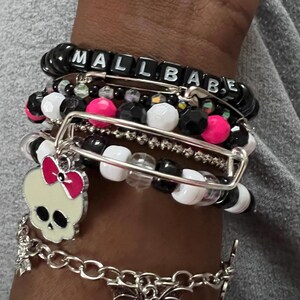 Op de afbeelding: Een stapel armbanden met zwarte en witte kralen, roze kralen en een zilveren bedel met een schedel en een roze strik. De bovenste armband heeft zwarte letterkralen die "MALL BABE" spellen.