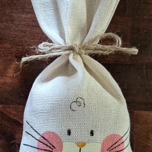 May include: A white fabric bag with a bunny face design. The bunny has pink cheeks, a yellow nose, and black whiskers. The bag is tied with twine.