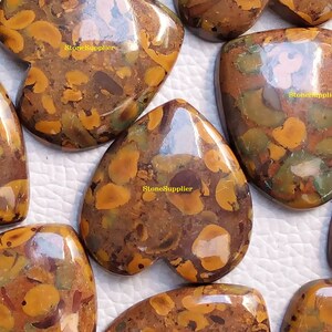 May include: A collection of seven heart-shaped, polished, brown, yellow and green jasper stones. The stones have a mottled, speckled pattern. The stones are arranged in a semi-circle on a white surface. The text "StoneSupplier" appears on three of the stones.
