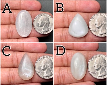 May include: Four white moonstone cabochons of varying shapes and sizes, each displayed next to a US quarter for scale. The cabochons are oval, teardrop, and triangular.