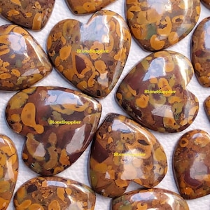 May include: A collection of heart-shaped, polished, brown and gold jasper cabochons with a mottled pattern. The cabochons are arranged in a random pattern on a white surface.
