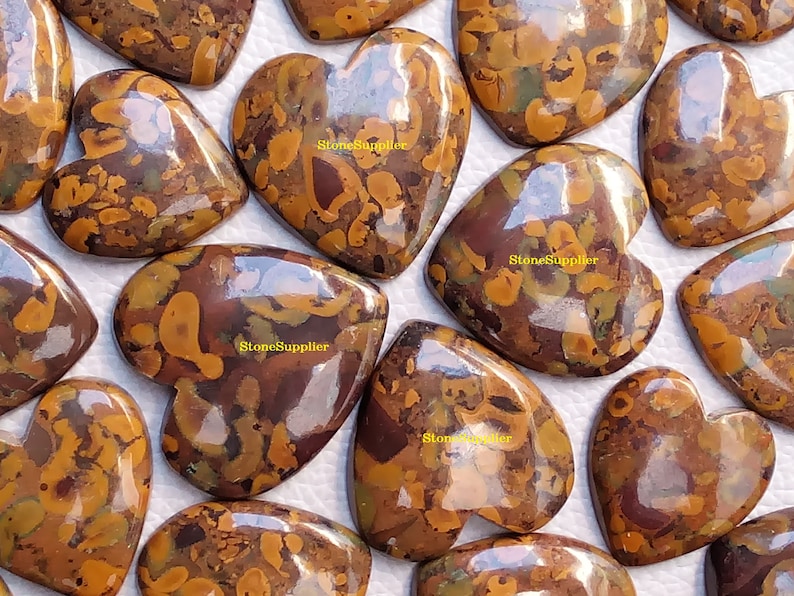 May include: A collection of heart-shaped, polished, brown and gold jasper cabochons with a mottled pattern. The cabochons are arranged in a random pattern on a white surface.