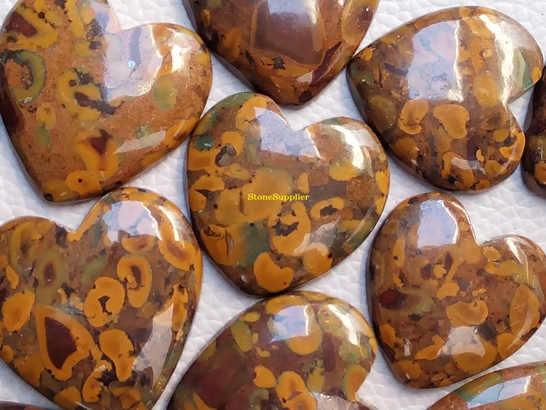 May include: A collection of eight heart-shaped polished stones with a brown, yellow, and green marbled pattern. The stones are arranged in a semi-circle on a white surface. The text "StoneSupplier" is visible in the centre of the image.