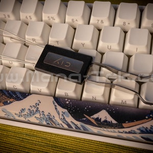 May include: A black keyboard keycap removal tool with a silver metal handle. The tool is lying on a white keyboard with a blue and white keyboard wrist rest featuring a Japanese wave design.