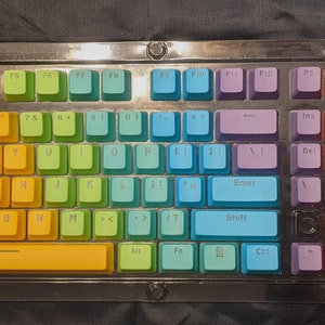 May include: A mechanical keyboard with a rainbow gradient keycap set. The keys transition from purple to red, orange, yellow, green, and blue. The keyboard has a clear frame with silver hardware and the text "Waves Variant" in the top left corner.