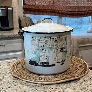 May include: A white enamelware picnic pot with a black handle and lid. The pot has a vintage graphic of a picnic scene and the text "Picnic Set That's Different". It sits on a round woven placemat.