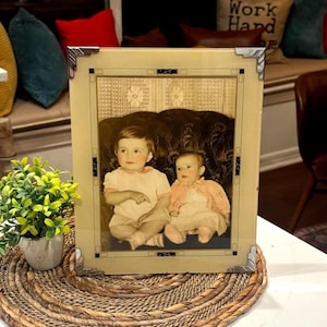 May include: A vintage framed photograph featuring two children in light-colored clothing. The frame is a light beige with silver corner accents. The photograph is displayed on a woven placemat with a small potted plant in the foreground.