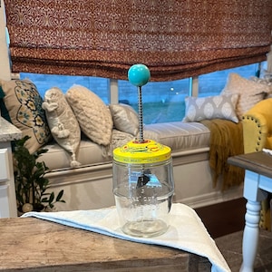 May include: A vintage yellow and green glass food mill with a turquoise knob and a metal crank. The mill has a glass jar with markings for measuring nuts, meats, and onions. The mill is sitting on a wooden surface.