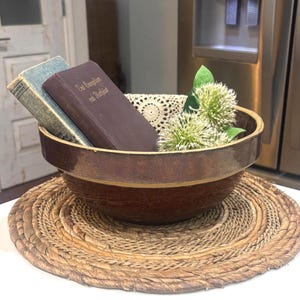 May include: A large, round, two-toned brown ceramic mixing bowl with a smooth, dark brown glazed rim and a textured, reddish-brown base. Inside the bowl are two vintage books, a cream-colored crocheted doily, and two white globe-shaped flowers.