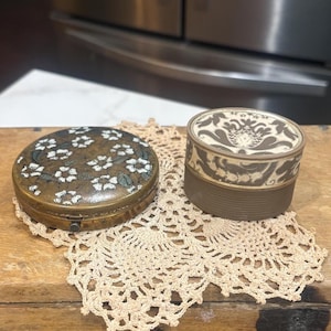 May include: Two antique decorative boxes. One is a round, brass-toned box with a floral design. The other is a small, cylindrical box with a patterned top and a brown base. Both are on a cream-colored doily.
