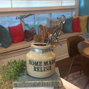 May include: A beige ceramic jar with the words "HOME MADE RELISH" in black lettering. The jar holds various kitchen utensils, including wooden spoons and a metal egg beater. The jar sits on a stack of old books.
