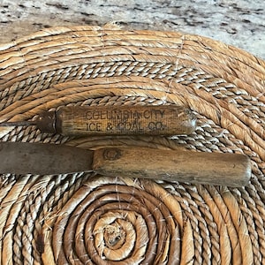 May include: Two antique tools with wooden handles and metal blades rest on a woven mat. One tool is a small ice pick, and the other is a small knife. The ice pick has the text "COLUMBIA CITY ICE & COAL CO."