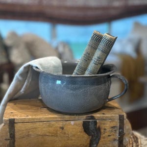 May include: A gray metal chamber pot with a handle, sitting on a wooden box. Two vintage books with gold lettering are resting on the rim of the pot. The books are titled "The Secret Red Barn Farm" and "The Mystery of Lilac Inn".