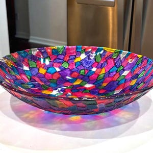 May include: A colorful glass bowl with a mosaic design. The bowl features a vibrant pattern of geometric shapes in shades of purple, pink, green, yellow, and blue. The bowl is round and sits on a white surface.