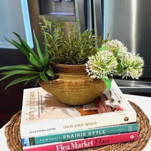May include: A decorative arrangement featuring a textured, light brown ceramic bowl filled with green plants and white, spiky flowers. The bowl rests on a stack of books with the titles "own your space", "Prairie Style", and "Flea Market Flair".