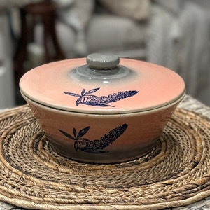 May include: A pink and gray ceramic serving bowl with a lid. The bowl has a blue and white floral design with a stylized flower and leaves. The bowl is sitting on a woven placemat.