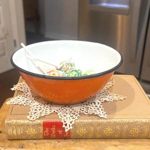 May include: A bright orange enamel bowl with a white interior and a thin black rim. The bowl contains several lollipops with white sticks and colorful wrappers. It rests on a beige crocheted doily placed atop a vintage book with gold lettering.