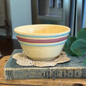 May include: A vintage yellow ceramic bowl with horizontal stripes in light blue and maroon. The bowl is sitting on a white lace doily and a blue book with the title "The Mystery at the Ski Jump".