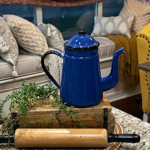 May include: A cobalt blue enamelware coffee pot with a black handle and lid. A wooden rolling pin with black handles rests in front of the pot. The pot sits on a wooden box with greenery, on a countertop.