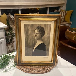 May include: A framed black and white portrait of a woman in a dark dress. The portrait is in a gold-toned frame with a double mat. The frame rests on a woven placemat on a white surface.