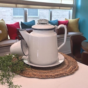 May include: White enamelware coffee pot with a glass lid and a matching cup. The pot has a black trim and a handle. The cup is hanging on the side of the pot. The set is on a white plate and a woven placemat.