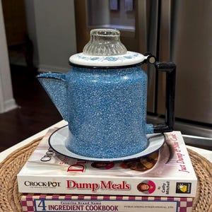 May include: A blue speckled enamel coffee pot with a glass top and black handle sits atop a stack of cookbooks. The cookbooks include "Crock-Pot Dump Meals" and "4 Ingredient Cookbook". The coffee pot is on a white plate.