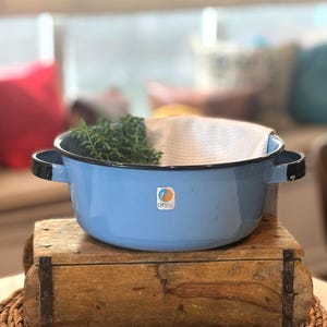 May include: A light blue enamelware pot with black handles and rim, resting on a wooden box. The pot contains a white cloth and green herbs. A logo is visible on the side of the pot. The pot is a vintage kitchenware item.