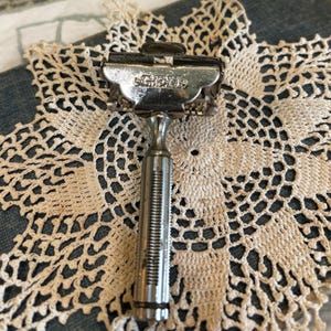 May include: A silver-toned metal safety razor featuring a textured cylindrical handle and a hinged head mechanism. The word Gillette is embossed on the top metal plate of the razor head, which is designed to hold a double-edged blade securely.