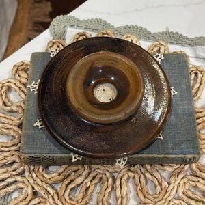 May include: A circular, dark brown glazed ceramic candle holder with a wide, flat rim and a central recessed cup featuring a small circular opening at the base. The surface of the pottery displays a glossy, mottled finish with variations in tone.