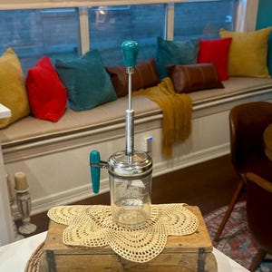 May include: A vintage glass and chrome butter churn with a teal handle and knob. The churn is sitting on a crocheted doily on a wooden box.