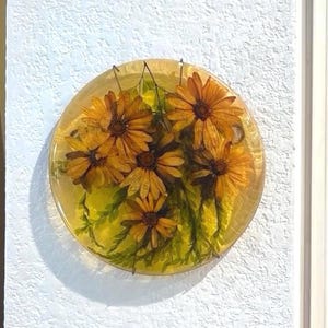 May include: A circular, translucent yellow resin wall plaque featuring embedded pressed orange and yellow flowers with green stems and leaves. The piece has a smooth, glossy finish and displays a botanical arrangement of five distinct blossoms.