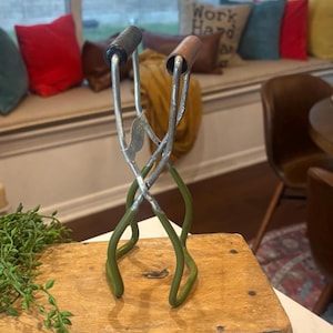May include: Vintage metal jar lifter with green handles and black and brown grips. The tool is designed to lift hot jars from boiling water. It is sitting on a wooden box.