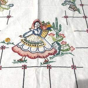 May include: A white fabric tablecloth featuring embroidered designs of a person carrying a basket of fruit, green cacti, and small flowers. The fabric is decorated with a black grid pattern accented by small red crosses at each intersection point.