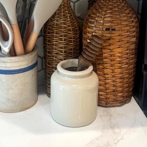 May include: A cream-colored ceramic jar with a wooden utensil inside, set on a white countertop. Behind it are two woven wicker containers and a stoneware crock with a blue stripe. Kitchen decor.