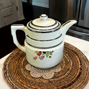 May include: A cream-colored ceramic teapot featuring a stepped lid and body with silver-toned metallic trim. The side of the teapot is decorated with a colorful floral pattern consisting of red, orange, purple, and green tulip-like flowers.