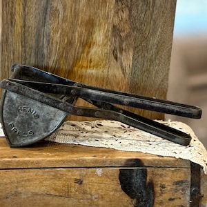 May include: An antique metal potato ricer with the text "KENIS PRESS" stamped on the head. The ricer is resting on a wooden box with a lace doily. The background includes a wooden plank and a blurred interior.