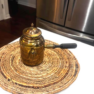 May include: A cylindrical brass coffee pot with a polished metallic finish, featuring a removable lid with a small finial knob, a short spout, and a straight black handle extending horizontally from the side of the main vessel body.