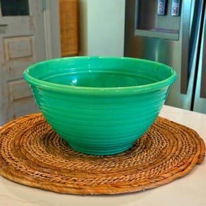 May include: A vibrant green ceramic bowl with a ribbed exterior sits atop a round, woven placemat. The bowl's bright color contrasts with the natural tones of the placemat, creating a visually appealing kitchenware item.