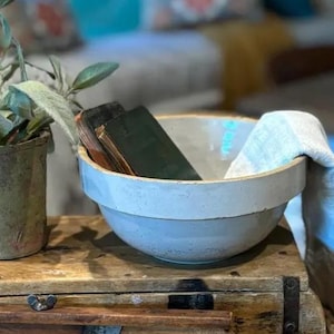 May include: A light blue ceramic bowl with a white rim, containing books and a folded linen towel. A small potted plant sits to the left on a wooden chest. The bowl is approximately 10 inches in diameter.