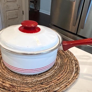 May include: A white enameled metal saucepan with a matching lid, featuring a bright red handle and a red knob on the lid. The base of the pot is decorated with three thin, horizontal red stripes, and the rim is finished with a silver metal edge.