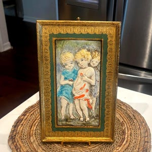 May include: A framed artwork featuring three figures in a classical style. The frame is gold with a green border. The figures are depicted in light colors, with one wearing a blue garment and the others in white, with orange accents.