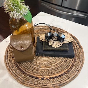 May include: A brown leather case with a button closure sits on a woven, circular placemat. A pair of small, black binoculars rests on a stack of dark books and a lace doily. A brown glass vase with white flowers is also present.