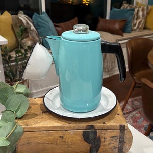 May include: A vintage turquoise enamelware coffee pot with a black handle and a clear glass lid. The coffee pot sits on a white plate with a black rim. A white and blue enamelware cup is attached to the pot.