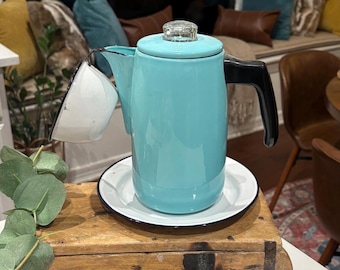 May include: A vintage turquoise enamelware coffee pot with a black handle and a clear glass lid. The coffee pot sits on a white plate with a black rim. A white and blue enamelware cup is attached to the pot.