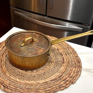 May include: A small, antique brass saucepan with a lid and long handle, resting on a woven placemat. The pot has a tarnished, aged appearance, suggesting it is vintage. The handle and lid handle are also brass.