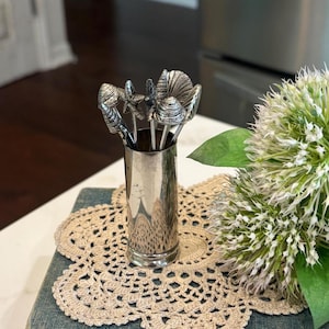 May include: A set of silver-toned metal cocktail picks featuring various seashell and starfish finials, housed in a matching cylindrical metal holder. The picks are arranged upright within the container, showcasing detailed textures on each nautical topper.