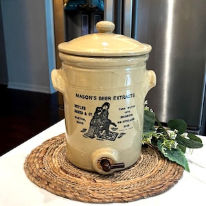 May include: A vintage beige stoneware beer extract dispenser with a lid, handles, and a spigot. The front features a black and white graphic and text that reads "MASON'S BEER EXTRACTS, BUTLER BREED & CO, BOSTON, MASS". It sits on a woven placemat.
