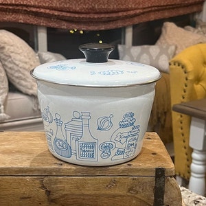 May include: White enamelware pot with a black lid and blue illustrations of cooking ingredients, including garlic, onions, oil, and herbs. The pot is sitting on a wooden surface.